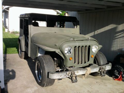 Olive green Jeep CJ-5 with a black soft top, seen from the front at a slight angle under a carport