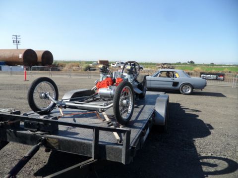 1973 Ford Ranchero - A dragster chassis is loaded onto a trailer, with a gray coupe parked nearby under a clear sky.