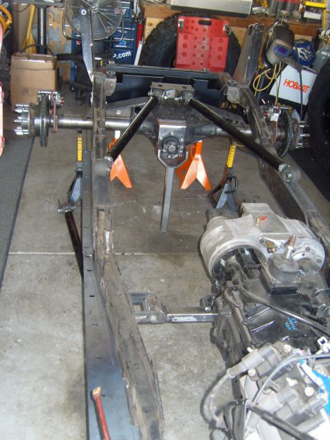 1985 Jeep CJ7 - The frame of a Jeep CJ is visible, supported by jack stands, with drivetrain components attached.