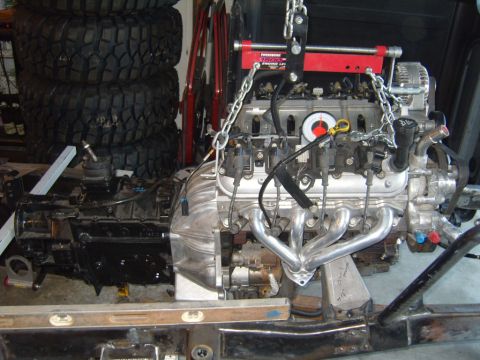 1985 Jeep CJ7 - Engine and transmission assembly being worked on, suspended by a hoist with exhaust headers visible.