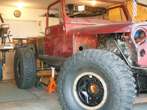 1985 Jeep CJ7 - A red Jeep CJ-series is undergoing modifications in a garage, supported by jack stands with large tires.