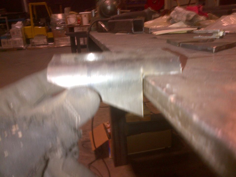 Metal bracket held by a gloved hand, with a workbench and forklift in the background