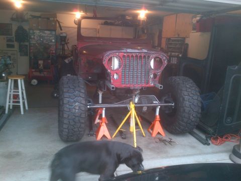 1985 Jeep CJ7 - A red Jeep CJ is elevated on jack stands in a garage, showing its front end and large tires.