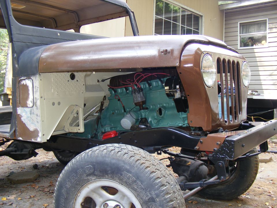 Front view of a Jeep CJ-5 with its engine exposed, showing the teal-colored engine block and brown hood