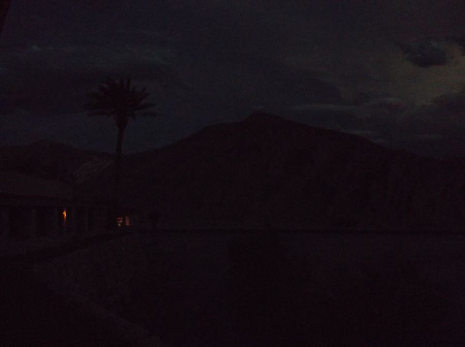 Dark desert landscape with mountains, a palm tree, and a building with lights on