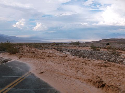 2009 Kenworth T800 - A desert road is partially submerged by floodwaters and debris, with mountains visible in the distance under a dramatic sky.