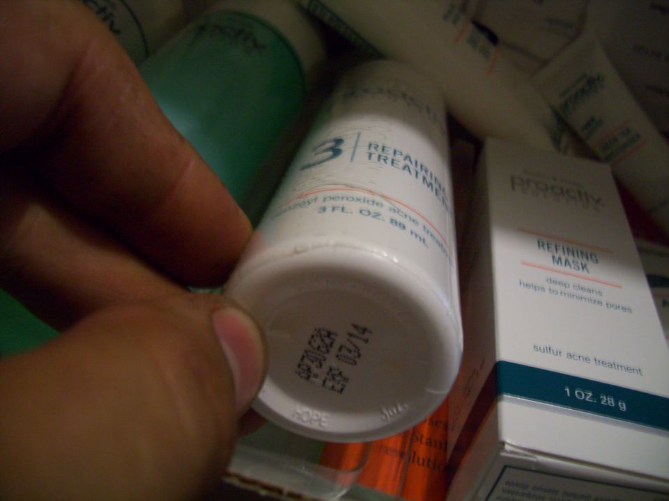 Hand holding a Proactiv Repairing Treatment bottle, with other Proactiv products visible. Expiration date 03/14 stamped on bottle