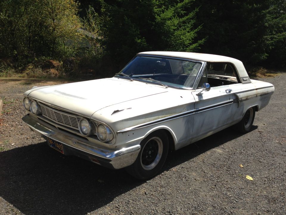 White 1964 Ford Fairlane coupe with primer patches on the body, viewed from the front three-quarter angle