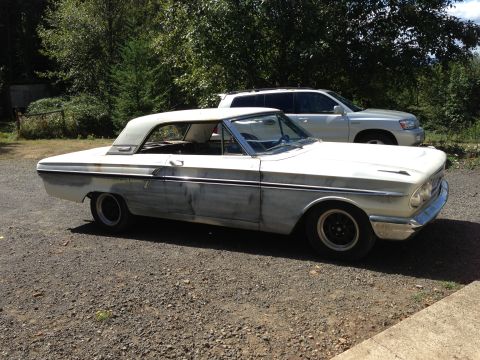 1964 Ford Fairlane - A white 1964 Ford Fairlane coupe, showing primer patches on its body, sits parked outdoors next to a modern SUV.
