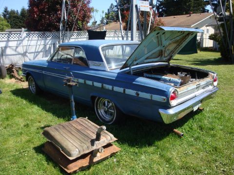 1964 Ford Fairlane - A blue 1964 Ford Fairlane sedan sits in a grassy yard with its trunk open, revealing disassembled parts.