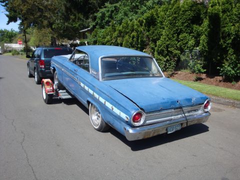 1964 Ford Fairlane - A blue 1964 Ford Fairlane coupe is seen from the rear, being towed on a trailer down a residential street.