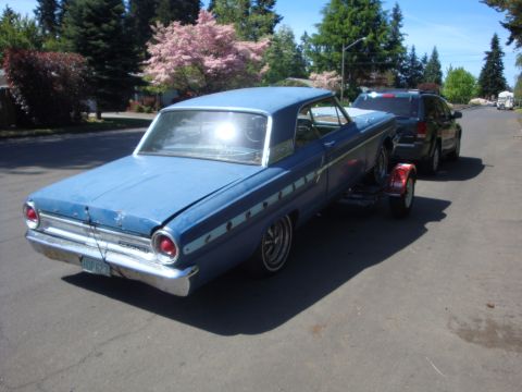 1964 Ford Fairlane - Blue 1964 Ford Fairlane is being towed on a trailer by a black SUV on a sunny day.