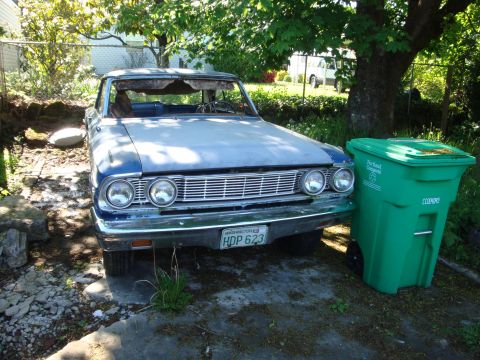 1964 Ford Fairlane - A 1964 Ford Fairlane sits parked next to a green trash bin, showing its front grille and headlights.