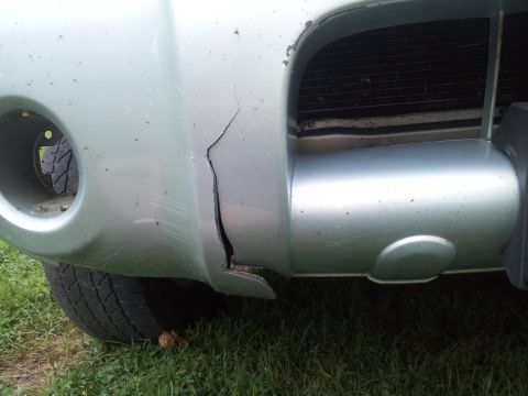 2003 Nissan Frontier - A cracked bumper on a silver truck is visible, with grass and a tire in the foreground.