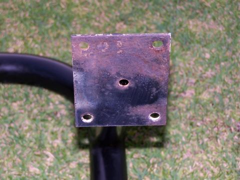 1973 Ford Bronco - A rusty square metal mounting plate with four holes is attached to a black curved metal tube. The plate is positioned on a background of gr…