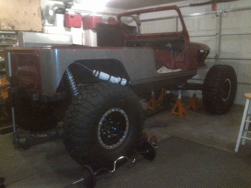Jeep CJ body on jack stands in garage, missing doors and roof. Large off-road tires are visible
