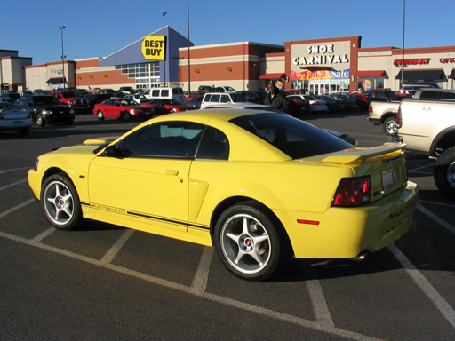 Yellow 2003 Ford Mustang parked in a lot with stores in the background