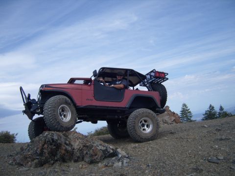 1985 Jeep CJ7 - A red Jeep CJ-series navigates rocky terrain under a partly cloudy sky.
