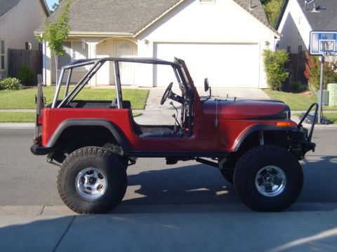 1985 Jeep CJ7 - This red Jeep CJ7, modified with a roll cage and off-road tires, is parked on a suburban street.