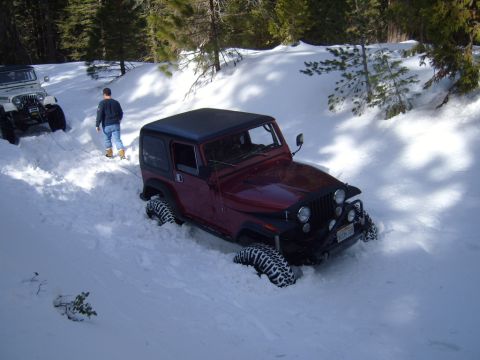 1985 Jeep CJ7 - Two Jeeps are stuck in deep snow on a wooded trail, with one vehicle attempting to pull the other out.