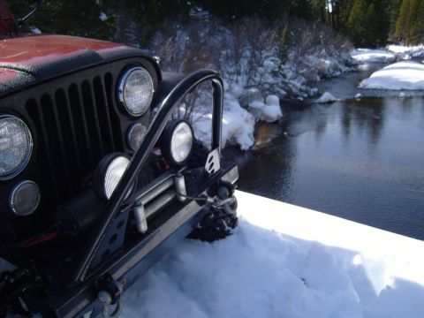 1985 Jeep CJ7 - A Jeep CJ is positioned in the snow near a cold river, with trees in the background.