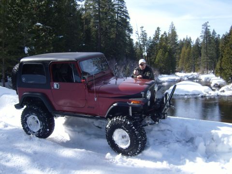 1985 Jeep CJ7 - A man leans on the hood of a maroon Jeep CJ7 parked in the snow next to a partially frozen river.