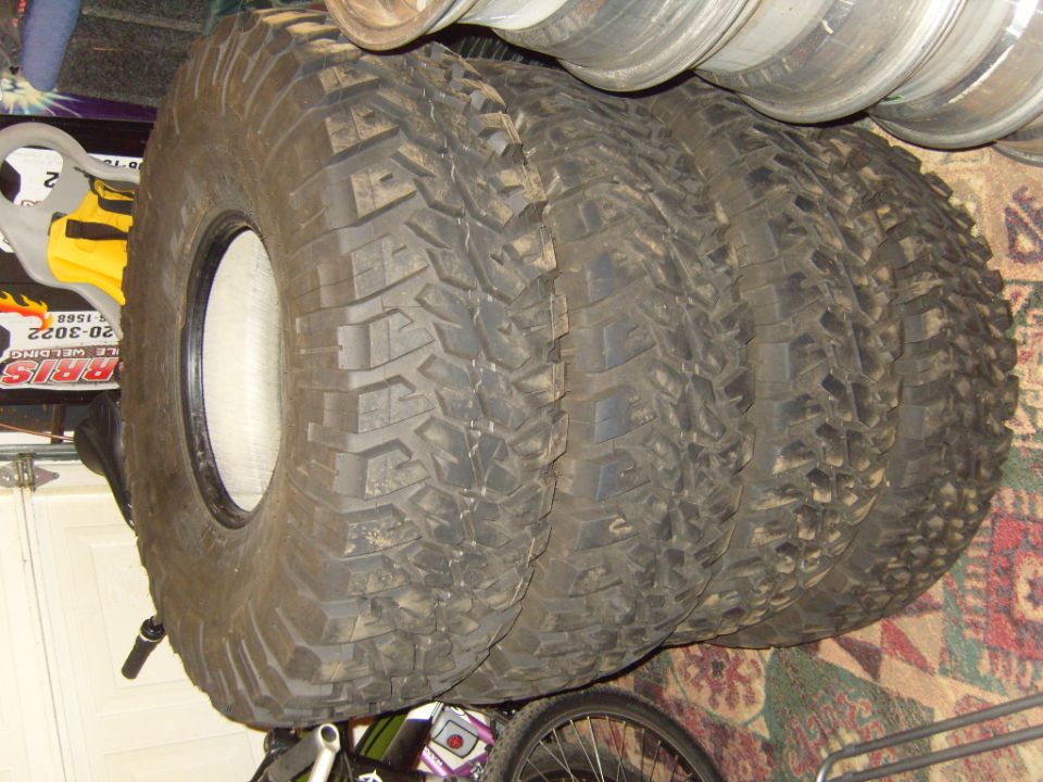 Four off-road tires stacked, one with a white rim, on a carpeted floor