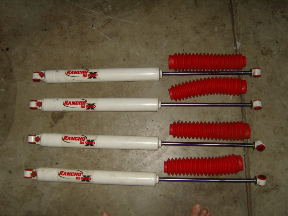 Four white Rancho RS9000 shock absorbers with red bellows laid out on a concrete surface