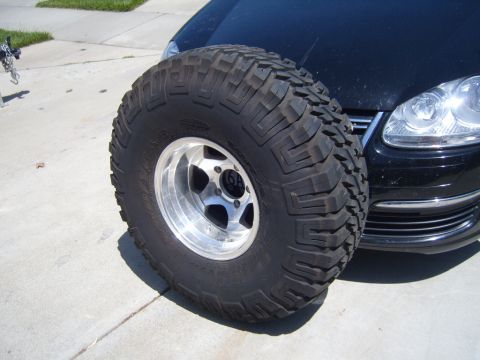 1985 Jeep CJ7 - A large off-road tire is positioned in front of the bumper and headlight of a black car.