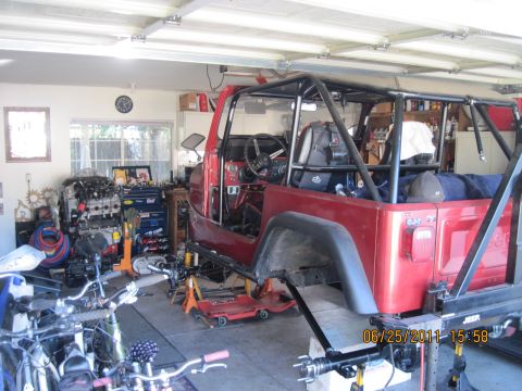 1985 Jeep CJ7 - Red Jeep CJ-series body with roll cage, on jack stands in a garage, with bicycles and an engine nearby.