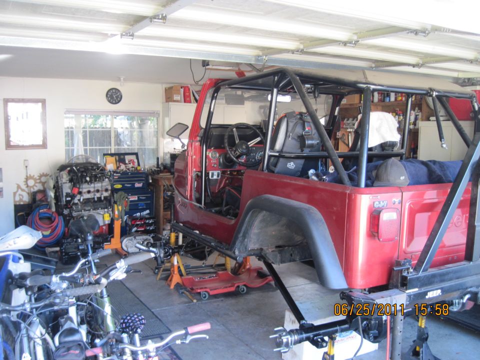 Red Jeep CJ-series body with roll cage, missing doors and fenders, on jack stands in a garage