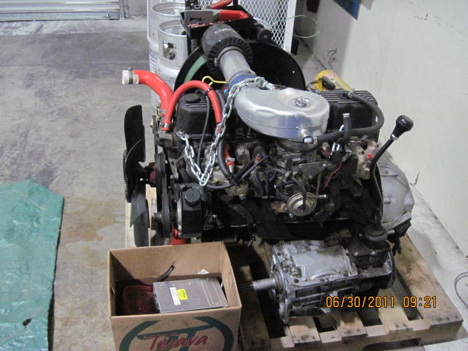Engine and transmission assembly on a wooden pallet, with a box of parts nearby. Date stamp reads 06/30/2011