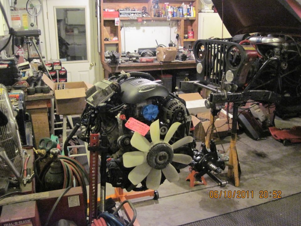 Jeep CJ-series grille and engine on stands in a garage. The grille is black with round headlights. A VORTEC engine is in the foreground