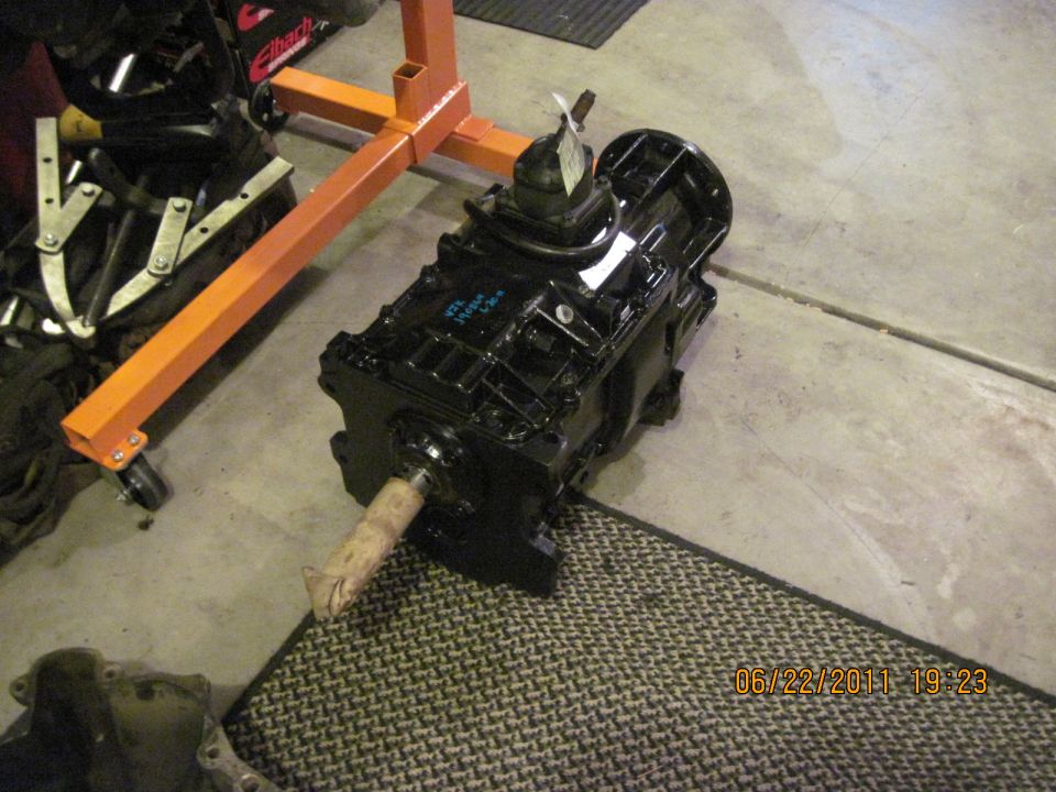 Black transmission with a tag on top, sitting on a mat next to an engine stand