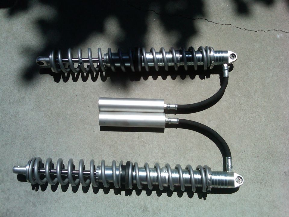 Two silver shock absorbers with black hoses and reservoirs sit on a concrete surface