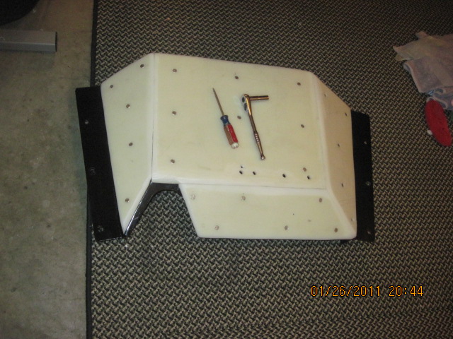 White fiberglass automotive part with black trim, on a patterned carpet. A screwdriver and ratchet are on top