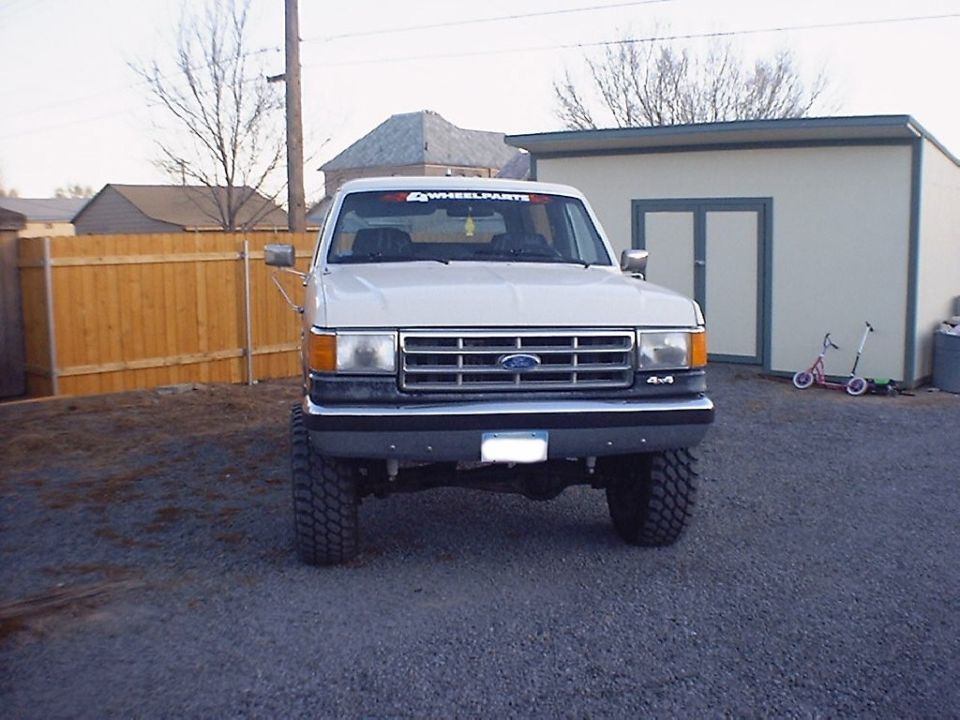 Front view of a lifted tan Ford 4x4 with 4WheelParts windshield banner and tall mud tires on gravel
