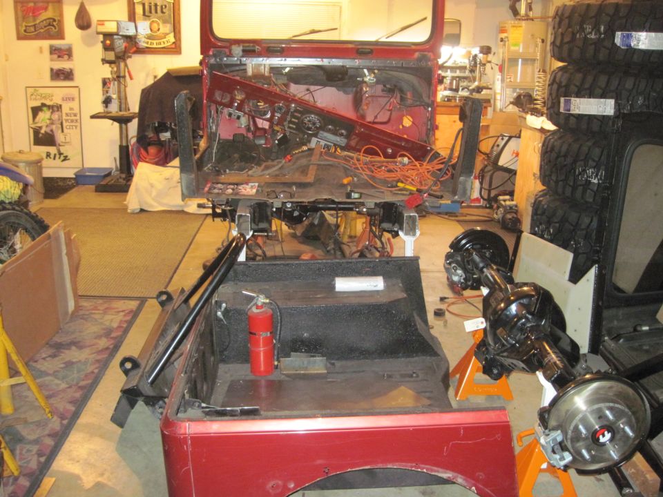 1985 Jeep CJ7 disassembled in a garage, showing the interior and chassis. A rear axle assembly is on an orange stand
