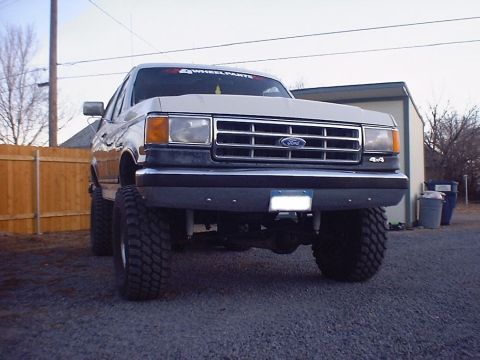 1988 Ford Bronco - Low front view of a lifted two-tone Ford 4x4 with a white upper body and dark lower body, sitting on aggressive mud-terrain tires and showi…