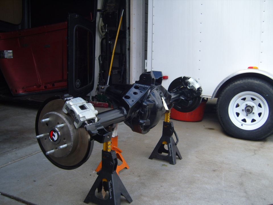 Jeep rear axle assembly with disc brakes on jack stands, red Jeep body in background, white trailer wheel to the right