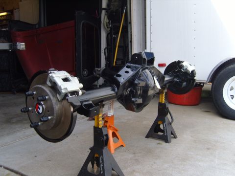 1985 Jeep CJ7 - A rear axle assembly, likely from a Jeep, is supported by jack stands in front of a red vehicle and a trailer.