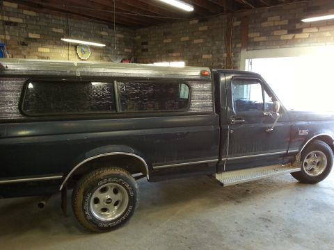 1989 Ford F-150 - A dark blue pickup truck with a camper shell is parked inside a garage.