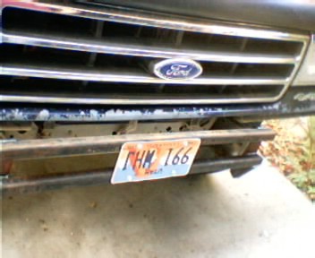 1991 Ford Bronco - Close-up of the front grille of a dark Ford truck showing the blue oval Ford emblem, horizontal grille bars, and a rusty steel bumper below…