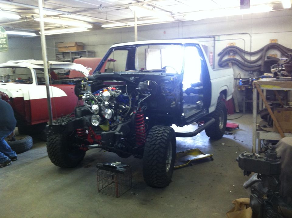 White 1980s Ford Bronco chassis with engine exposed, lifted with large tires