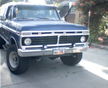 1991 Ford Bronco - Dark blue 1970s-era Ford pickup on a residential driveway, shot low from the front three-quarter angle. It has block FORD lettering across…