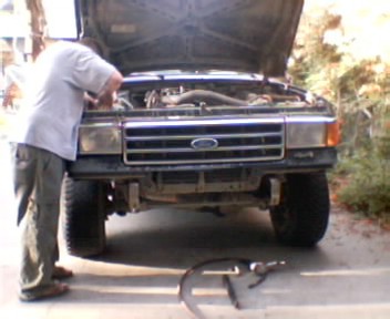 1991 Ford Bronco - A person in a gray shirt leans under the raised hood of a dark late-1980s Ford truck parked in a driveway, with the front bumper removed to…