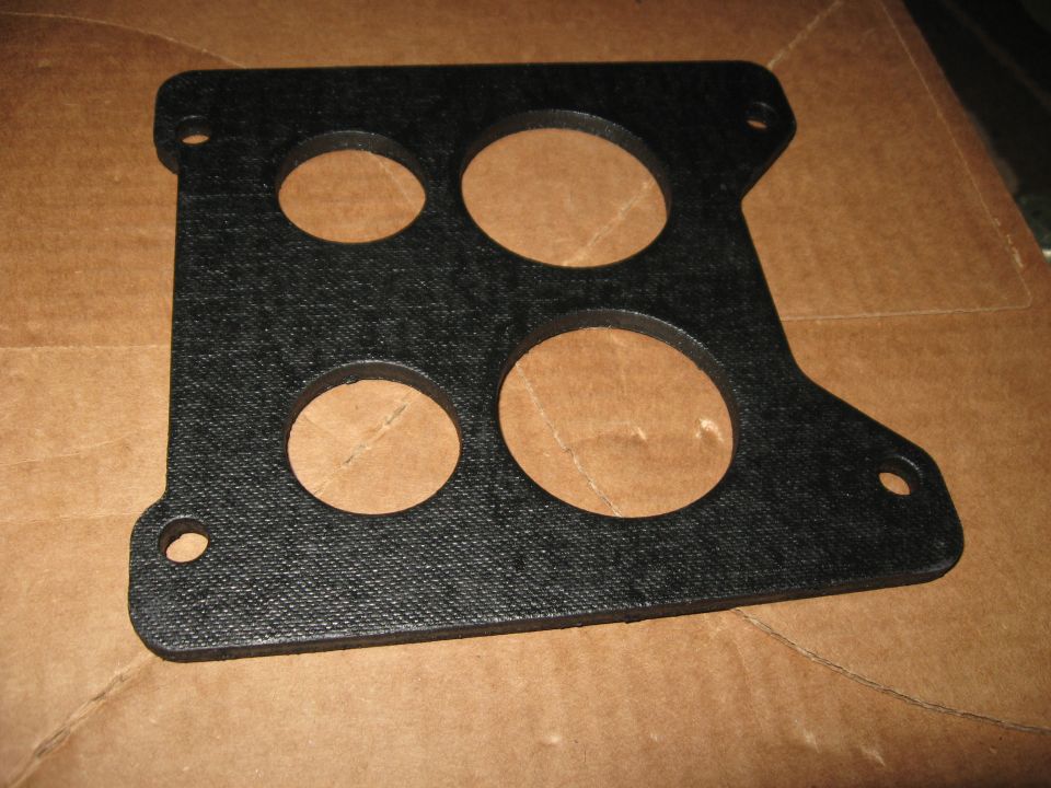 Black carburetor gasket with four circular openings on a cardboard surface