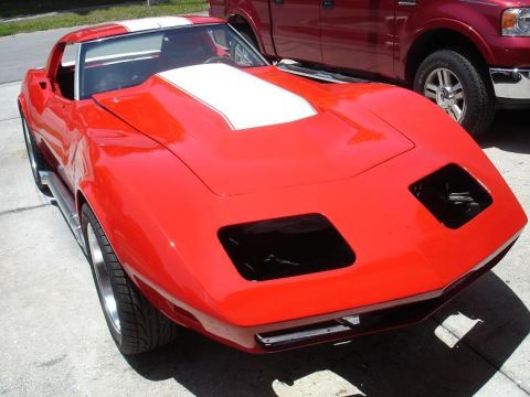 1976 Chevrolet Corvette - A red Chevrolet Corvette with white racing stripes is parked next to a red pickup truck, missing its pop-up headlights.