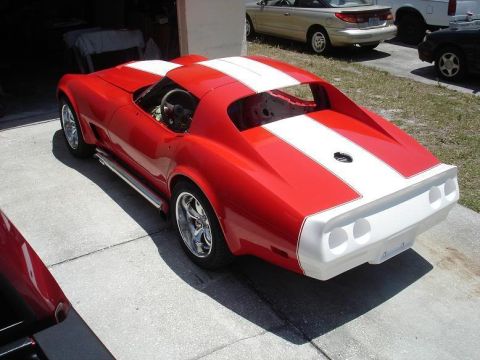 1976 Chevrolet Corvette - A red Chevrolet Corvette with white racing stripes is parked on a concrete driveway, with other vehicles visible in the background.