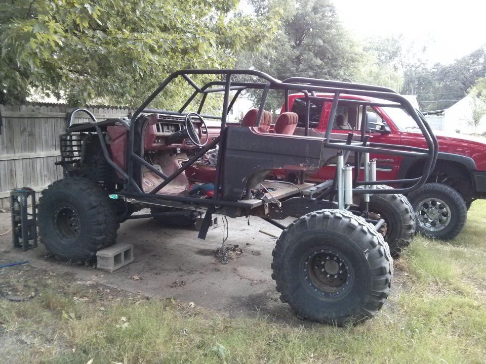 Modified off-road vehicle with roll cage, large tires, and red interior, parked next to a red pickup truck